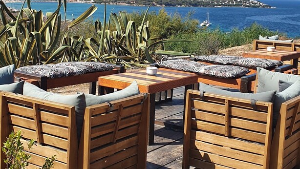 Une terrasse en bois, aménagée avec du mobilier d’extérieur, notamment des canapés et une table, qui offre une vue dégagée sur la côte, la mer et le paysage lointain.