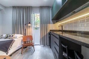 A modern hotel room with a bed, a desk, a chair, a kitchen area with a stove, and a view of greenery outside. - Seng 106 in S o Paulo (São Paulo)