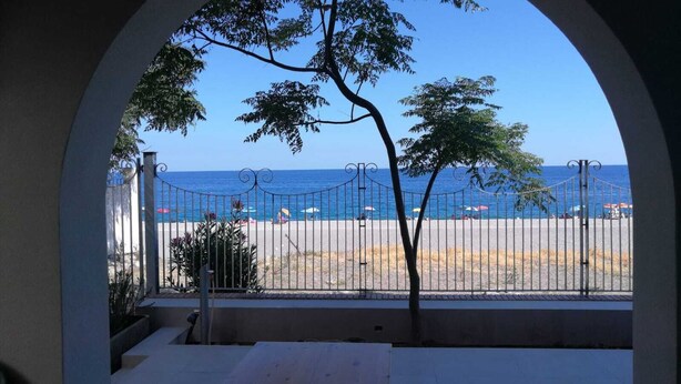 A view through an arched window of a beach with a metal fence, a tree, and a clear blue sky. - Villa by the sea, perfect for families (Mascali)