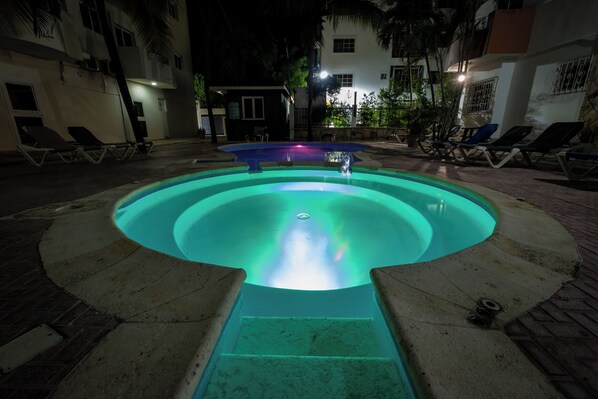 Una piscina con una fuente central, rodeada de sillas de descanso y un pequeño edificio.