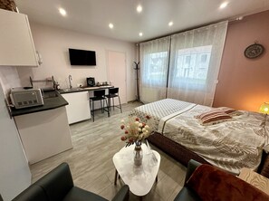 A compact hotel room with a kitchenette, a bed with decorative pillows, a small dining table, and a vase with flowers. - Studio Millésime (Nogent-l'Artaud)