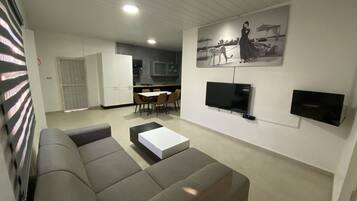 A modern living room with a grey sectional sofa, a dining area with a table and chairs, a kitchen in the background, and a mounted TV on the wall.