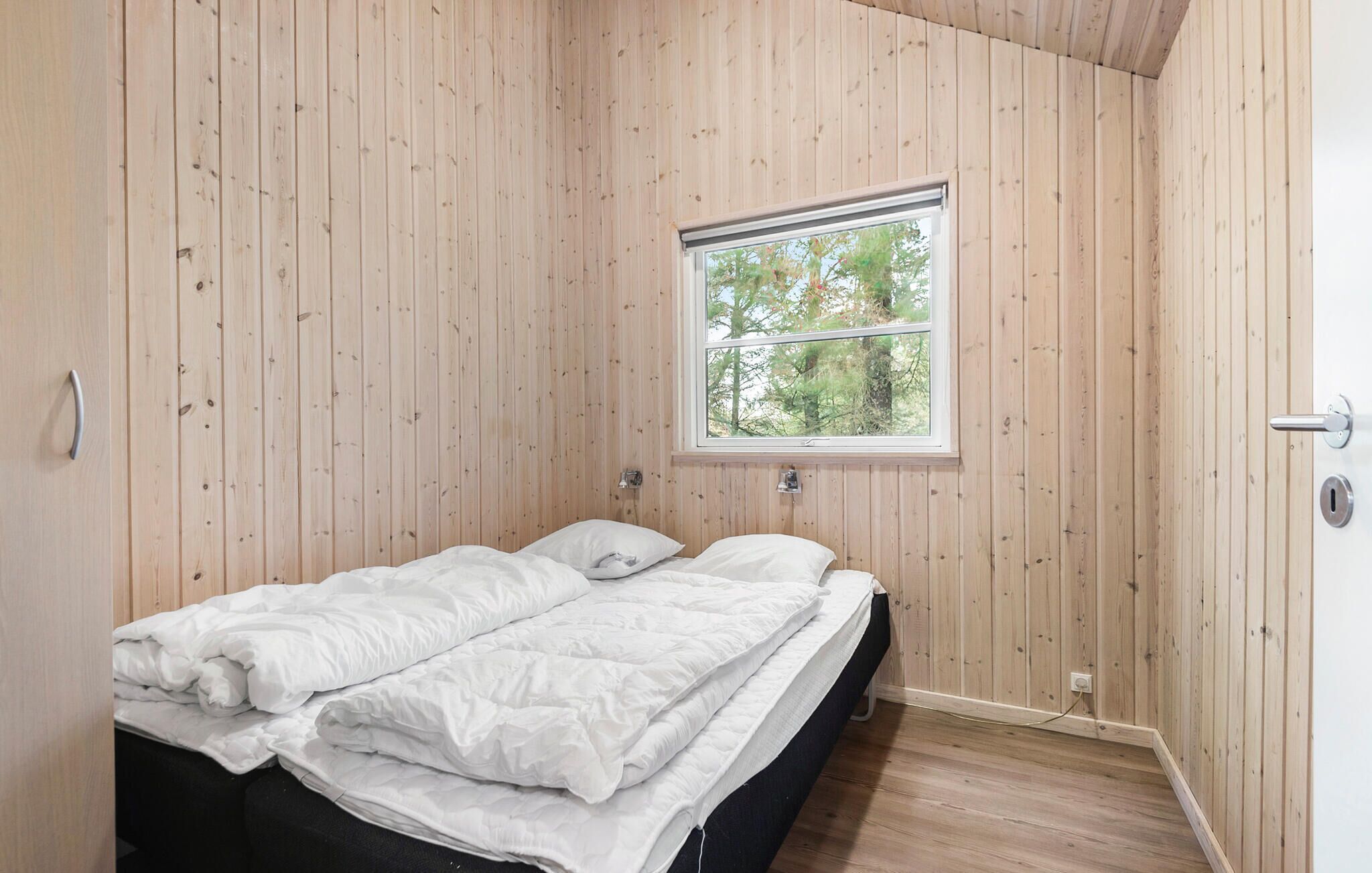 Una camera da letto con parete rivestita in legno, una finestra vista alberi, un letto con biancheria bianca e una porta con occhiello.