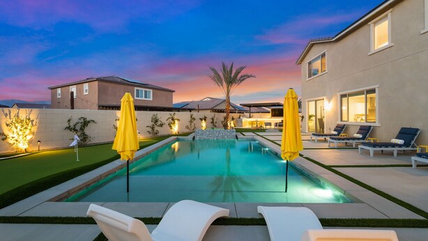 Piscina com espreguiçadeiras e guarda-sóis, cercada por casas e paisagismo bem iluminado.