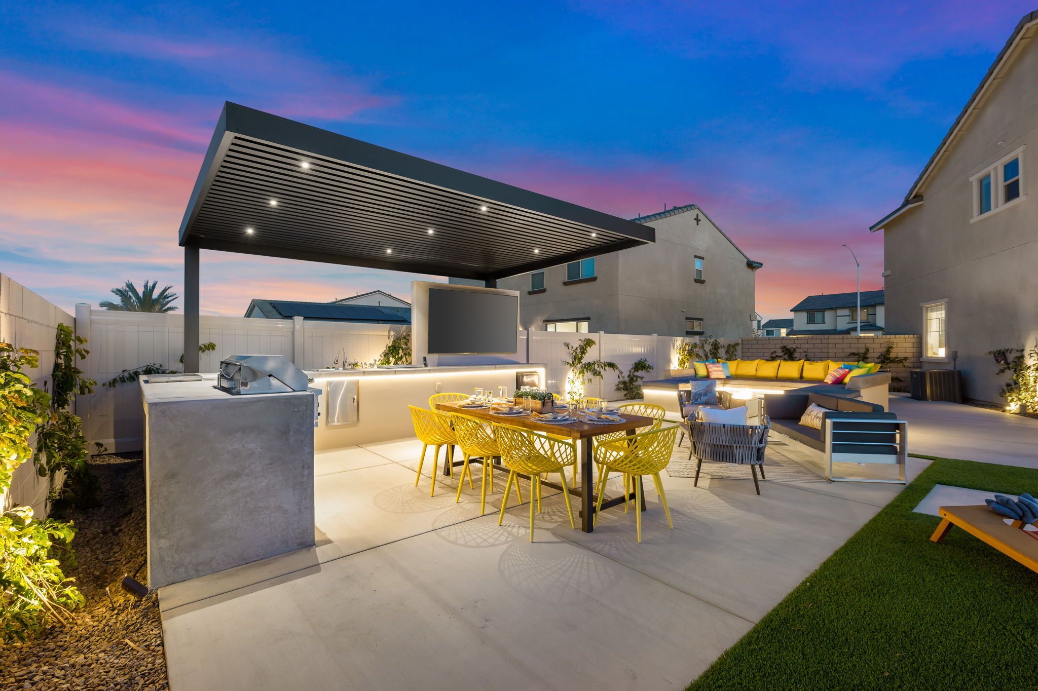 A modern outdoor kitchen area with a built-in grill, a dining table with yellow chairs, and a covered seating area with cushions.