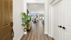 Um corredor com piso de madeira, paredes brancas e um vaso com planta. Há uma porta à esquerda e uma porta branca à direita.