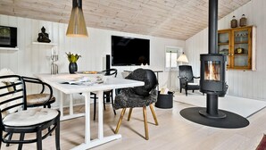 A cozy living room with a wooden ceiling, a black wood-burning stove, a dining table with chairs, and a TV.
