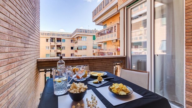 Varanda com mesa posta para refeição, incluindo pipoca, copos e uma garrafa, com vista para um prédio residencial.
