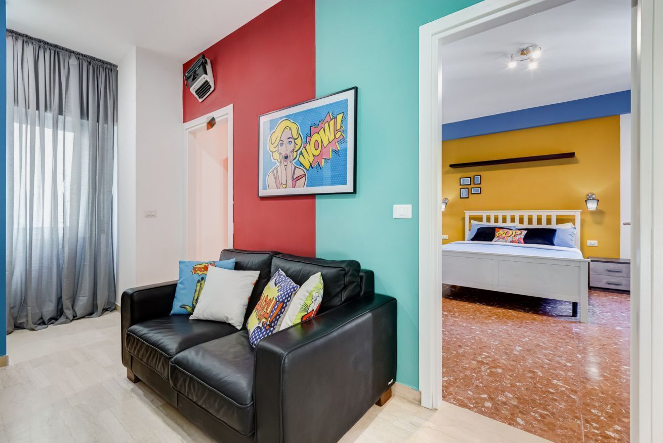 A room with a black leather sofa, a bed with a white frame, a colorful wall, and a red and blue partition.