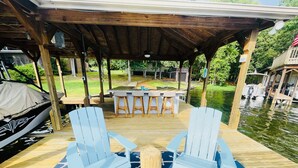 Een overdekt terras buiten met een houten tafel en stoelen, een overdekte barbecue en uitzicht op een vijver.