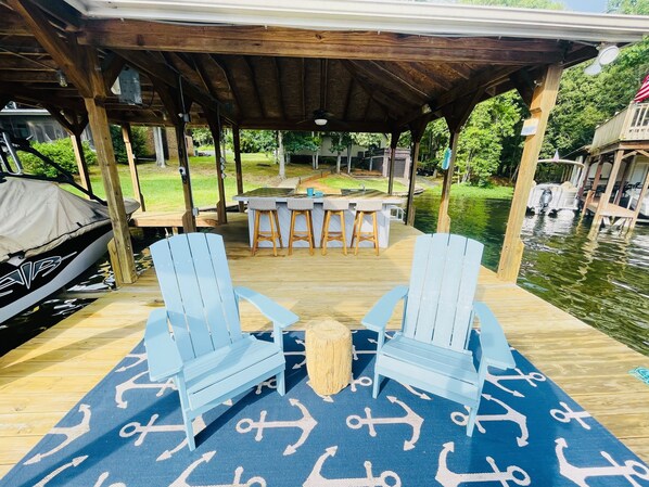 A covered outdoor deck with a wooden table and chairs, a covered grill, and a view of a pond.