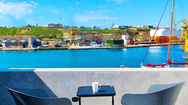A seaside view with a clear sky, a dock with a sailboat, and a terrace with chairs and a table. - Savynomad Harbour Residences Bormla (Cospicua)