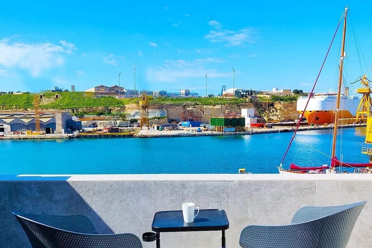 A seaside view with a clear sky, a dock with a sailboat, and a terrace with chairs and a table.