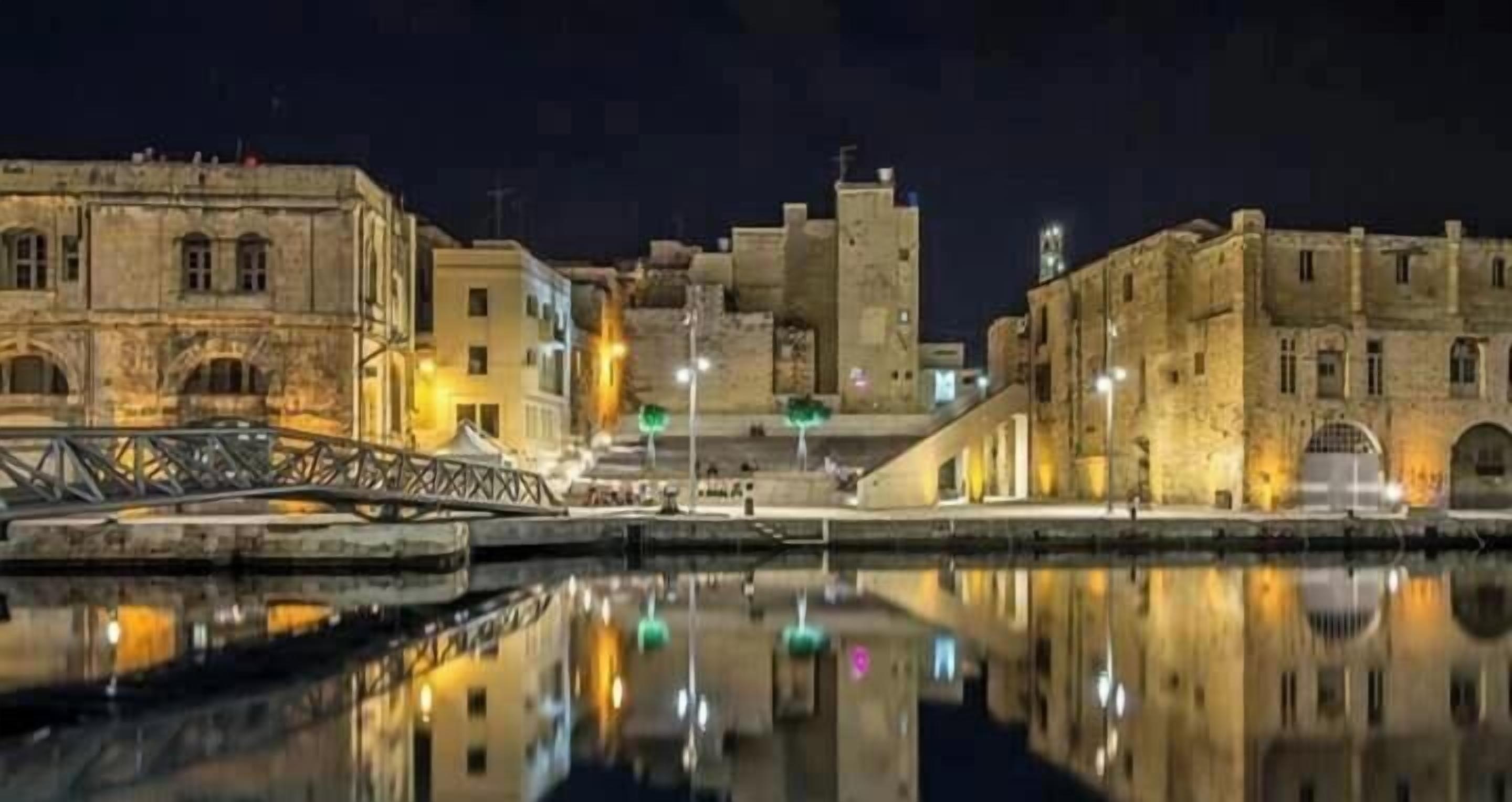 Une vue nocturne d’un quartier historique situé au bord de l’eau, avec ses bâtiments en pierre, un pont et une rivière paisible qui reflète les lumières.