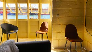 A room with a large window overlooking a construction site, two chairs, and a small table. - Savynomad Harbour Residences Bormla (Cospicua)