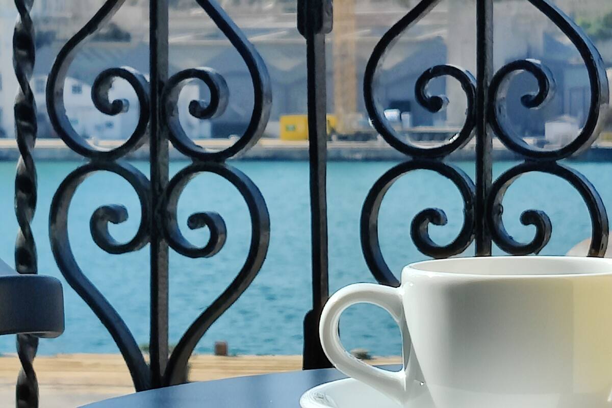 A white cup on a saucer on a table, with a metal railing and a view of water in the background.