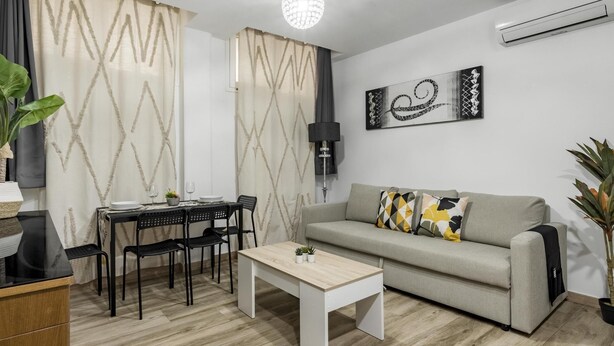 Een moderne woonkamer met een eethoek, een grijze bank met patroon kussens, een houten salontafel en een kroonluchter.