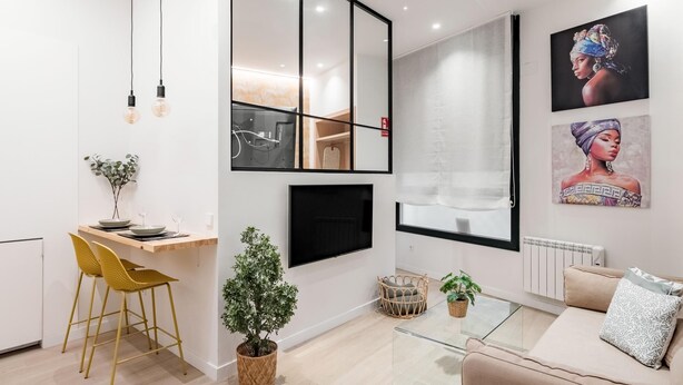 Un salon moderne avec un canapé, une table basse en verre, un coin repas avec des chaises jaunes, une petite plante et des œuvres d’art accrochées au mur.