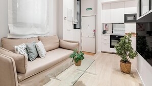 Une chambre d’hôtel moderne avec un canapé beige, une table basse en verre, une plante en pot et un coin cuisine.