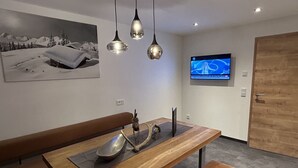 A modern dining area with a wooden table, bench seating, pendant lighting, a wall-mounted TV, and a framed picture.