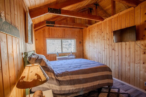 A wooden cabin room with a bed, a desk, a chair, a window, and a mounted television.