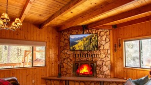 Interior acogedor de una cabaña con paredes de madera, chimenea de piedra, televisor montado y ventana con vista a un bosque.