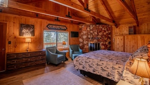 Un dormitorio tipo cabaña de madera con cama, dos sillas, tocador, chimenea de piedra y ventana con vistas a árboles.
