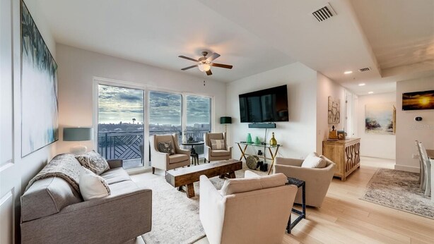 A spacious living room with a large window, a ceiling fan, and a flat-screen TV mounted on the wall. - Luxury new condo steps to the beach with Gulf Views, heated pool, & hot tub (Madeira Beach)