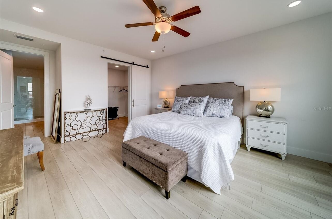 Een slaapkamer met een bed, een nachtkastje, een lamp, een plafondventilator, een bankje en een deur naar een andere kamer.