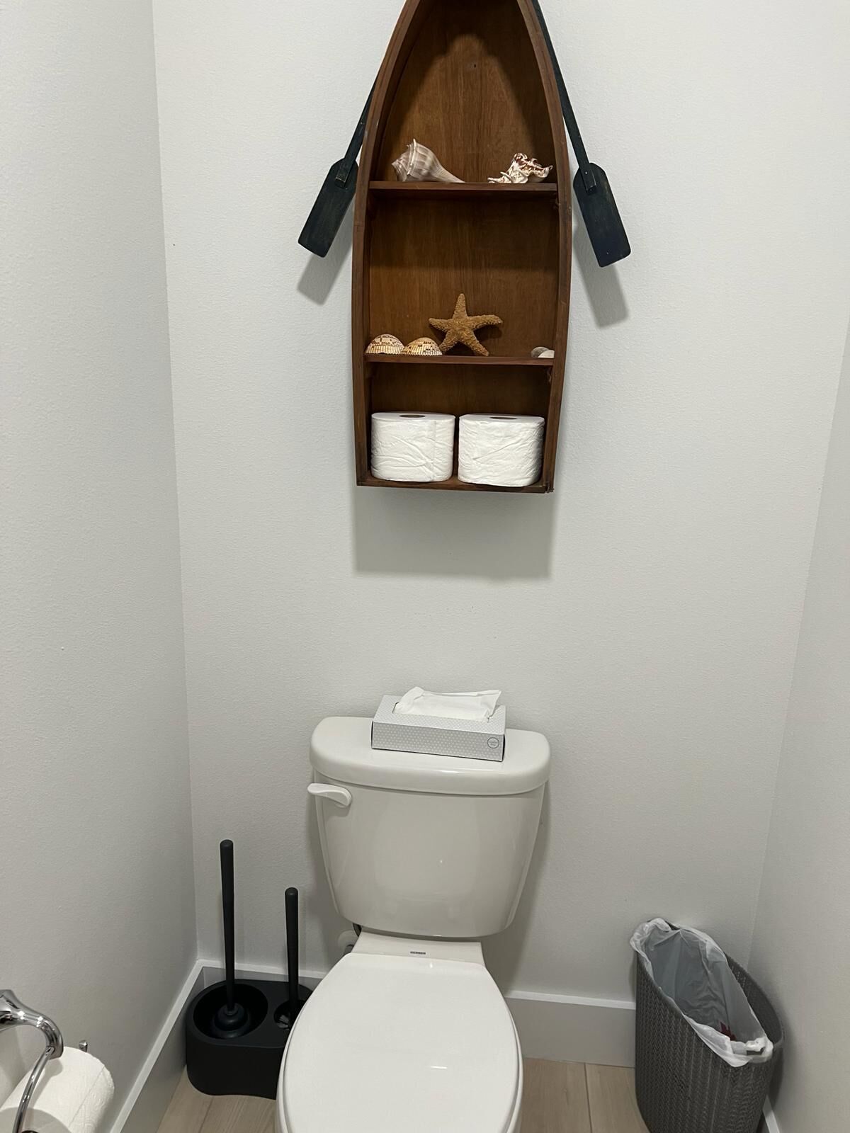 A bathroom with a toilet, a shelf above it holding a starfish and seashells, and a trash bin to the right.