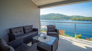 Un balcón con mobiliario de mimbre, una mesa de vidrio y vistas a un lago y colinas.