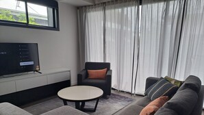 Een moderne woonkamer met een grijze bank, een ronde salontafel en een grijze fauteuil. Een flatscreen televisie is aan de muur bevestigd, en er is een raam met dunne gordijnen te zien.
