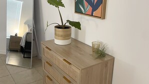 A wooden dresser with a plant in a white pot, a framed abstract painting, and a mirror reflecting a room with a bed and a window.