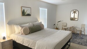 A bedroom with a bed, bedside tables, a ceiling fan, a window with blinds, a wall-mounted mirror, and a small side table with a candle.