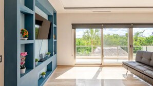 A modern living room with a sofa, a TV on a blue wall, and a balcony with a view of greenery. - Exclusive Tulum-Akumal Stay | Dock, Beach Club, Pool, Cenote & Golf (Akumal)
