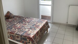 A bedroom with a patterned bedspread, a single pillow, a sliding glass door to a balcony, and a radiator on the wall. - Appartement T2 Tout Confort, Grand Balcon, Saint Nazaire Ouest (Saint-Nazaire)