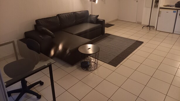 A living room with a black leather sofa, a glass table, and a small round table. There is a white door and a kitchen area in the background. - Appartement T2 Tout Confort, Grand Balcon, Saint Nazaire Ouest (Saint-Nazaire)
