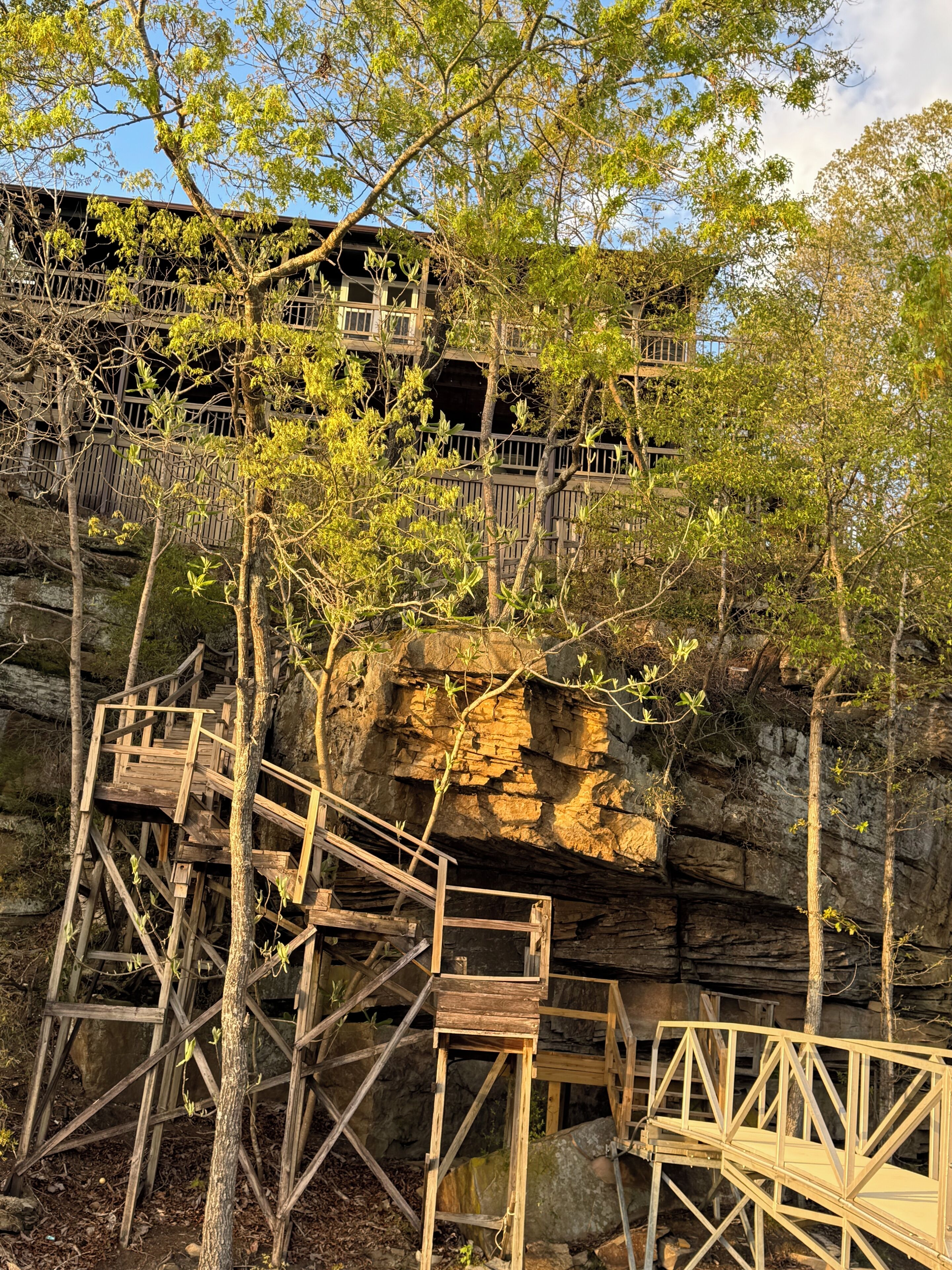 Wooden walkways and platforms built into a rocky cliffside, with trees and a building in the background.