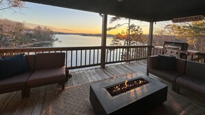 Eine überdachte Terrasse mit Feuerstelle, Gartenmöbeln und Blick auf einen See bei Sonnenuntergang.