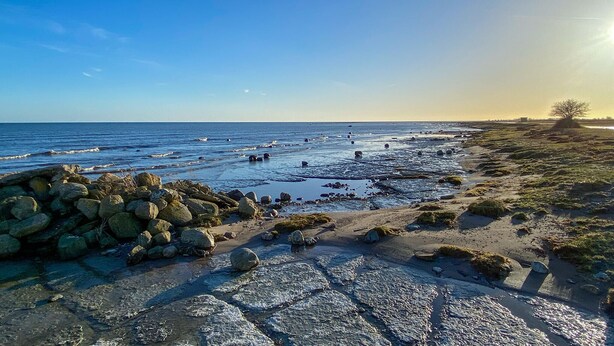 Una spiaggia rocciosa con le onde che si infrangono sulla riva, cielo sereno e un tramonto sullo sfondo.