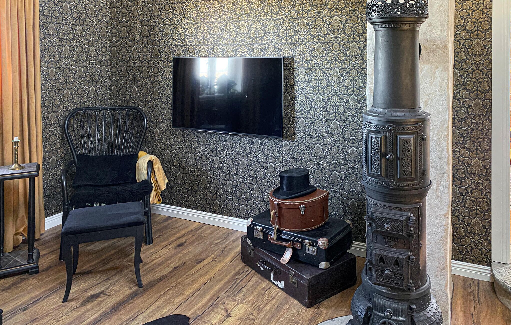 A room with a wooden floor, a black chair, a television mounted on the wall, and a decorative stove.