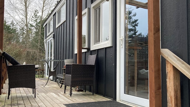 En overdækket terrasse med træmøbler og bænk, der fører hen til et hus med mørk facade og store vinduer.