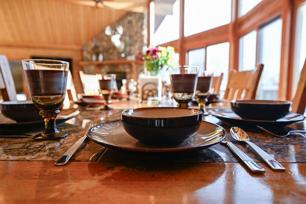Mesa de comedor preparada con platos, tazones y cubiertos en una habitación con paredes de madera y ventanas grandes.
