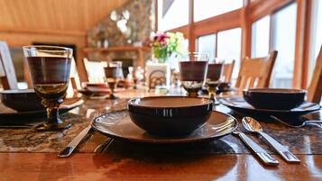 Mesa de comedor preparada con platos, tazones y cubiertos en una habitación con paredes de madera y ventanas grandes.