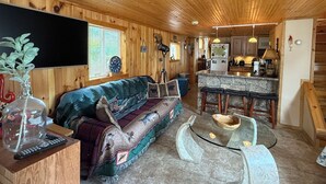 A cozy cabin interior with a wooden ceiling, a stone bar area, a sofa with a colorful blanket, and a dining table.