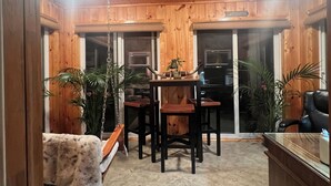 A cozy wooden cabin interior with a dining area featuring a wooden table and chairs, a bar area with a counter, and several potted plants.