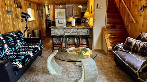 A cozy cabin interior with a stone bar, wooden walls, a black leather sofa, and a brown leather couch.