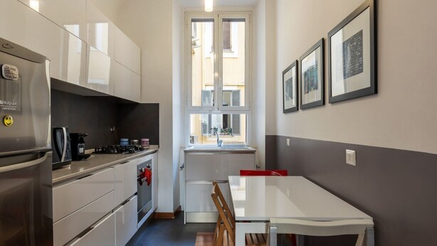 A compact kitchen with a refrigerator, stove, and sink, a dining table with chairs, and a window with a view of buildings. - GuestHost - Comfortable apartment located on the second floor, of six in total, of a building with a lift (NOT suitable for disabled people).In 60 m2 of space it comfortably accommodates three people, with a double bedroom and a single bedroom.Located in (Roma)