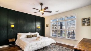 Un dormitorio con cama, ventilador de techo, ventana con persianas, mesita de noche y alfombra.