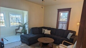 Ein Wohnzimmer mit einem Ecksofa, einem runden Couchtisch und zwei Sesseln. Es gibt ein großes Fenster mit Blick nach draußen.